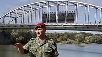 Ein Soldat hält draußen einen Vortrag. Im Hintergrund ein Fluss und eine Brücke.