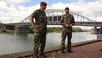 Zwei Soldaten stehen draußen im Gespräch. Im Hintergrund ein Fluss und eine Brücke.