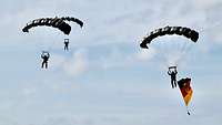 Drei Soldaten gleiten an Fallschirmen mit einer deutschen Nationalflagge unter bewölktem Himmel.