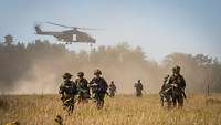 Soldaten laufen durch hohes Gras, im Hintergrund steigt ein Hubschrauber auf.