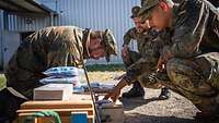 Drei Soldaten hocken am Boden, neben ihnen Holzkisten mit Munition.