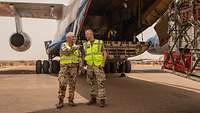 Zwei Soldaten mit Warnweste stehen hinter der geöffneten Laderampe eines Flugzeuges. 