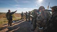 Ein Soldat steht vor zahlreichen Soldaten auf einem Berg bei Sonnenschein und spricht.
