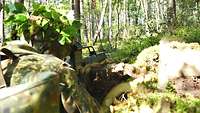 Ein Soldat liegt im grünen Wald mit Gewehr im Anschlag. Sein Helm ist mit Blättern getarnt.