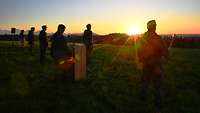 Soldaten stehen bei Sonnenuntergang vor einem Alpenpanorama auf einer Wiese angetreten.