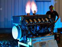 Tank engine Flames are shooting out of an engine in the dark. A man wearing ear protection is standing in the background.