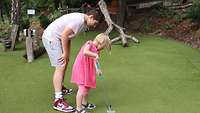 Ein Mädchen in einem rosa Kleid wird von einem größeren Jungen beim Minigolf angeleitet. 
