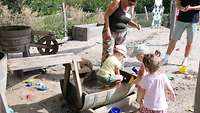 Kinder spielen im Sand und einer Wassertonne.