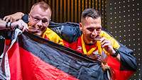 Zwei deutsche Sportler in Nationalflagge gehüllt, beißen auf ihre Bronzemedaillen und freuen sich. 