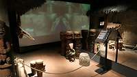 In einem dunklen Raum stehen Trommeln und Figuren aus Afrika, dahinter läuft ein Video. 