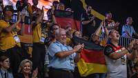 Menschen stehen auf einer Tribüne. Sie klatschen, schwenken deutsche Flaggen und feuern an.