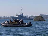 Ein graues Schiff, zwei Schlauchboote mit mehreren Personen und ein Fallschirm schwimmen im Wasser.