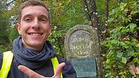 Ein Mann zeigt vor der Quelle der Ruhr in Winterberg mit erhobenem Daumen und Zeigefinger eine 2