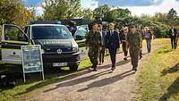 Zwei Soldaten gehen mit einem Mann in einem blauen Jacket an einem Fahrzeug der Feldjäger vorbei. 