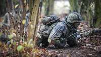 Ein Soldat kriecht durch das Gelände. Weitere Soldaten unscharf im Hintergrund.