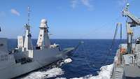 Zwei graue Schiffe fahren nebeneinander her. Auf beiden stehen jeweils mehrere Personen auf dem Oberdeck.