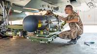 Ein Soldat kniet unter einem Eurofighter. Ein zylinderförmiger Behälter wird mit einer Hebebühne ans Flugzeug angebracht.