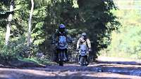 Zwei Soldaten fahren versetzt hintereinander auf einem dunklen Waldweg auf Motorrädern.