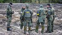 Sechs Soldaten stehen im schlammigen Gelände. Sie sprechen miteinander.