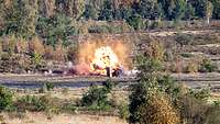 Ein Geschoss eines Kampfpanzers zerschlägt ein Hartziel im Gelände, ein Feuerball steigt auf.