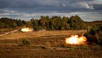 Zwei Kampfpanzer stehen auf einer Freifläche. Sie feuern mit ihren Rohren, ein Feuerball entsteht.