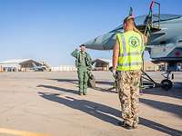 Zwei Soldaten stehen sich gegenüber und grüßen sich militärisch. Im Hintergrund sind Eurofighter in Sheltern zu sehen.