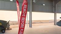Zwei große rote Anhänger sind zu sehen. Auf ihnen steht „Remove before Flight“. Im Hintergrund sieht man den Shelterbereich.