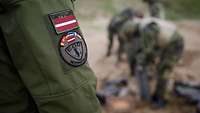 Patch am Ärmel eines lettischen Soldaten, im Hintergrund Soldaten auf einem Sprengplatz
