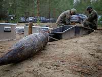 Ein großes altes Geschoss liegt auf Sandboden, im Hintergrund arbeiten zwei Soldaten.