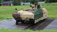 A dummy vehicle covered with camouflage nets is standing on a round wooden board.