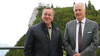 Two men in suits are standing in front of an alpine backdrop.