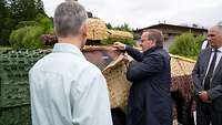 “MMT” on a dummy vehicle A man is looking at green and beige padded cloth nets. Two men are standing next to him.