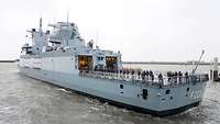 Ein Schiff der Marine fährt aus einem Hafen. Auf dem Heck stehen zahlreiche Soldaten.