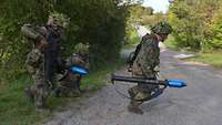 Ein Soldat kniet und sichert an einem Weg, zwei Kameraden mit Panzerfäusten überqueren diesen.