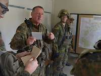 Ein Soldat mit einem Schreibblock informiert andere Soldaten, an einer Wand hängt eine Lagekarte.