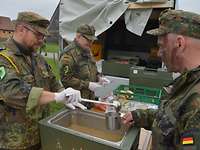 Der Kompaniefeldwebel gibt aus einer rechteckigen Therme an Soldaten eine heiße Suppe aus.