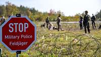 Im Vordergrund ein Schild „STOP Military Police Feldjäger“ und im Hintergrund Soldatinnen und Soldaten auf einer Wiese.