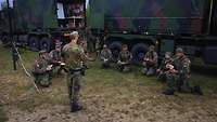 Neun Soldaten hocken im Halbkreis um einen stehenden Soldaten. Im Hintergrund sind zwei LKW.