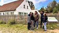 Eine Familie steht vor Bauernhaus, das umzäunt ist mit einem Holzzaun.