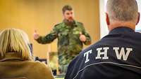 Ein Soldat spricht in ein Mikrofon. Das Publikum hört ihm zu.