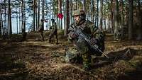 Ein Soldat kniet mit Waffe im Wald. Im Hintergrund laufen Soldaten mit roten Flaggen.