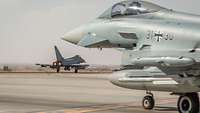 Im Vordergrund sieht man unscharf seitlich einen Eurofighter. Dahinter steht ein Eurofighter im Fokus der gerade startet.