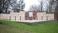 Ein Gedenkstein mit Gedenktafel vor einer Mauer mit vielen Namensfafeln im Wald der Erinnerung