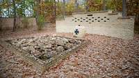 Ein Gedenkstein mit Eisernem Kreuz vor einer Mauer mit mehreren Tafeln im Wald der Erinnerung