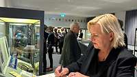 Eine Frau trägt sich in ein Gästebuch ein, im Hintergrund eine Glasvitrine