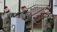 Ein Soldat steht am Rednerpult, zwei weitere links und rechts davon.