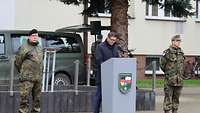 Ein Zivilist steht am Rednerpult, zwei weitere links und rechts davon.