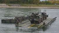 Eine Fähre schwimmt im Rhein, darauf ein Kran und ein Lkw. Sie wird von Booten angetrieben.