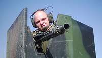 Ein Soldat mit Kopfhörern steht in einem gepanzerten Fahrzeugturm, aus dem ein Maschinengewehr ragt.