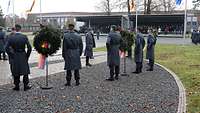 Soldaten stehen neben aufgebauten Kränzen, im Hintergrund wehende Flaggen und zwei Tribünen. 